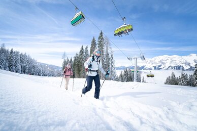 Zwei Skitourengeherinnen steigen durch den Schnee, während über ihnen ein Sessellift mit Skifahrern fährt. | © Josh Absenger