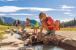 Two kids play at a water game station with channels and gates at Hopsiland Planai, mountains and trees in the background. | © Christine Höflehner