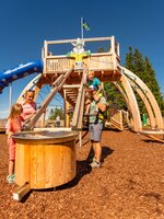 Familie spielt im Hopsiland Planai an Wasserrinne, im Hintergrund Holzspielstruktur mit blauer Rutsche und Hasenmotiv. | © Christine Höflehner