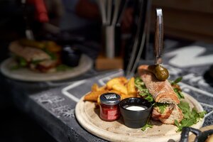 Ein knuspriges Baguette mit Speck, Rucola, Soßen, Essiggurke und Wedges wird auf einer Holzplatte im Sonnenlicht serviert. | © Dietmar Körbler