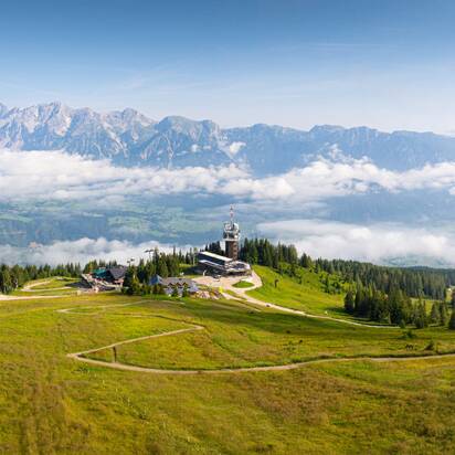Vast alpine pasture with a mountain station and observation tower, surrounded by meadows, forest, and mountain ranges. | © Josh Absenger