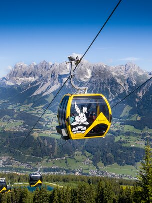 Gelbe Gondel mit Hopsiland-Motiv über der Planai mit Blick auf das Dachsteinmassiv und das grüne Ennstal im Sommer. | © Josh Absenger