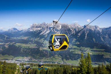 Yellow gondola with Hopsiland bunny above Planai with Dachstein mountains and green Enns valley in bright summer weather. | © Josh Absenger