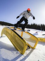 Snowboarder in a grey hoodie and orange beanie sliding on a yellow rail in Funpark Planai under bright sunshine. | © Roland Haschka QParks