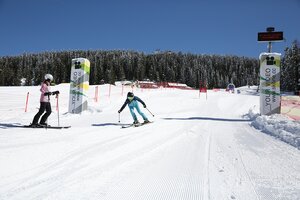 Skifahrerin in blauem Anzug erreicht die Ziellinie des Skimovie-Rennkurses auf der Planai, während eine Begleiterin zuschaut. | © Herbert Raffalt