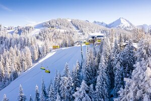 Aerial view of Planai with snowy trees, ski slopes, and a yellow cable car against a blue sky. | © Josh Absenger