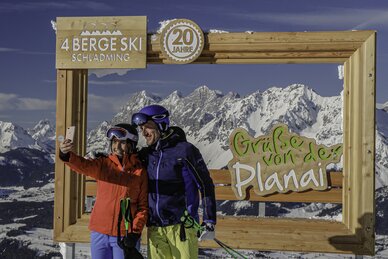 Zwei Skifahrer machen ein Selfie vor einem Holzrahmen mit verschneiten Bergen. | © Herbert Raffalt