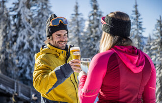 Two people clinking beer and wine glasses in front of a snowy winter landscape. | © Mirja Geh