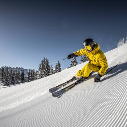 Ein Skifahrer in gelber Kleidung fährt rasant eine frisch präparierte Piste hinunter, während Schnee in die Luft wirbelt. | © Mirja Geh