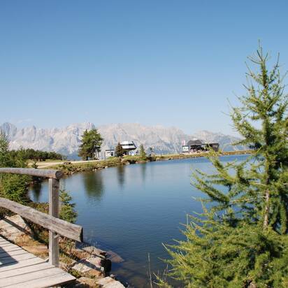 A boardwalk leads along the calm Reiteralm mountain lake, with Dachstein range and two cable car stations in sunny backdrop. | © Reiteralm Bergbahnen