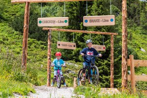 Two kids ride their mountain bikes into signed Junior Trails at Reiteralm, surrounded by wildflowers and green forest trees. | © Reiteralm Bergbahnen