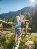 A boy joyfully balances on a log bridge by a stream, with family enjoying a sunny nature adventure on the Reiteralm mountain. | © Lorenz Masser