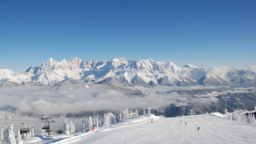 Blick auf verschneite Piste, Skifahrer und beeindruckendes Bergpanorama im Hintergrund.