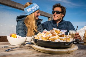 Kaiserschmarrn in Pfanne auf Holztisch, Mann und Frau im Gespräch, Reiteralm im Hintergrund. | © Lorenz Masser