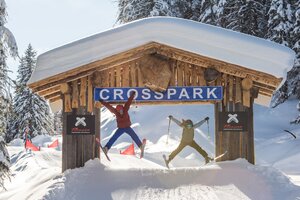 Zwei Skifahrer springen gleichzeitig unter einem Holzschild „Crosspark“ auf der Reiteralm. | © Lorenz Masser