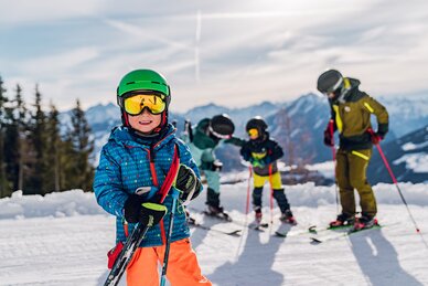 Ein Junge in Skiausrüstung steht grinsend auf der Piste und im Hintergrund sind die Eltern mit Geschwister.