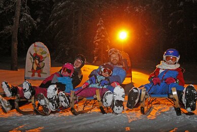 Family night sledding with helmets and headlamps on snowy track.