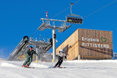 Two skiers descend in sunshine in front of Erlebnis Rittisberg and the chairlift station.