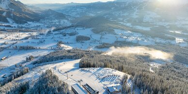 Drone shot of snowy Rittisberg with base station, slope and Enns Valley in sunlight.