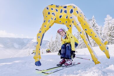 Skifahrerin mit gelber Jacke fährt durch eine gelbe Giraffenfigur, Schnee bedeckt, blauer Himmel, Berge im Hintergrund. | © Bergbahnen Filzmoos