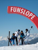 Eine Familie mit zwei Kindern steht unter einem roten halben Bogen auf dem Funslope steht und jeder hält einen Ski hoch in die Luft