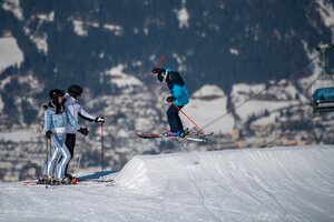 Skifahrer in blauer Ausrüstung springt im Beginnerpark vor winterlicher Kulisse.