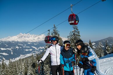 Familie in Skikleidung lacht auf der Piste mit Gondeln und Alpenpanorama im Hintergrund