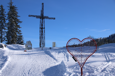Präparierter Schneeweg führt zu Kreuz aus Metall und ein rotes Herz aus Metall steht im Vordergrund