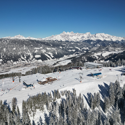 Eine Gondelstation, zwei Sesselliftstationen und eine Skihütte von oben und im Tal unten sind viele Häuser zu sehen