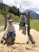 Zwei kleine Kinder spielen auf einer flachen Holzrutsche vor grüner Bergkulisse. | © Shuttleberg