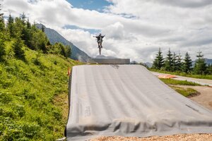 Mountainbiker springt mit ausgebreiteten Armen über Bagjump im Grünen vor Bergkulisse. | © Shuttleberg