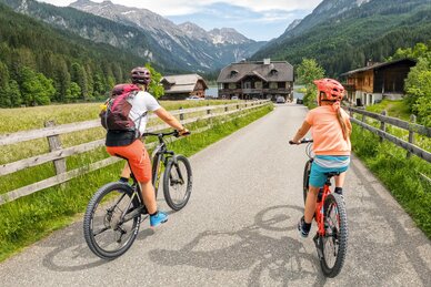 Adult and child ride mountain bikes through a green valley with wooden fences and mountains in the background. | © Shuttleberg