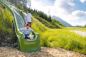 A toddler laughs while sliding down a green slide, an adult stands behind. | © Shuttleberg