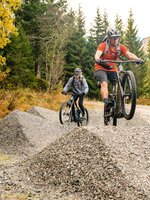 Two mountain bikers ride over gravel hills in the forest, one jumping. | © Shuttleberg