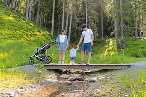 Parents with toddler and stroller cross a small wooden bridge in a green forest. | © Shuttleberg