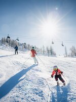 Kind und Erwachsene beim Skifahren auf sonniger Piste, Sessellift und Bäume im Hintergrund | © Shuttleberg