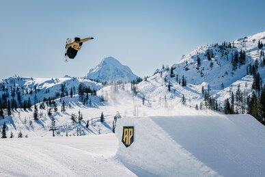Ein Snowboarder ist über eine Schanze aus Schnee gesprungen und macht in der Luft einen Grab | © MRochrbacher