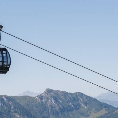 Two black panoramic gondolas float above green summer mountains, with snowy Alpine peaks in the far background. | © Snow Space Salzburg