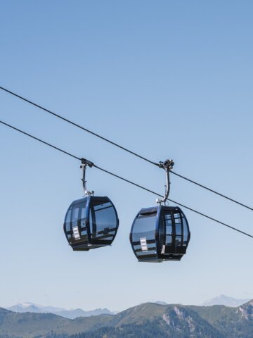 Zwei schwarze Panoramagondeln schweben über grünen Bergen im Sommer, im Hintergrund schneebedeckte Alpengipfel. | © Snow Space Salzburg