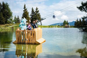 Familie mit Maskottchen auf Holzfloß in Bergsee, Gondel im Hintergrund, Bäume und Berge bei sonnigem Wetter sichtbar | © Snow Space Salzburg