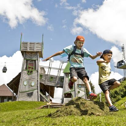 Two kids jump over a grassy mound in front of a fantasy play tower at Geisterberg adventure park in St. Johann, Salzburg | © sanktjohann.com | Mirja Geh