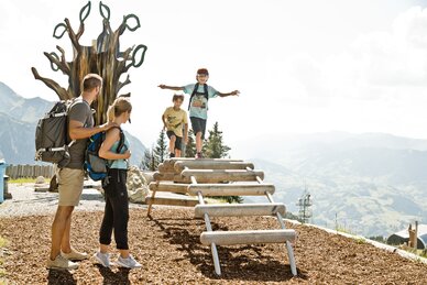 Two kids on wooden climbing frame, parents watching them, background shows mountains, trees and a large wooden sculpture | © sanktjohann.com, Mirja Geh