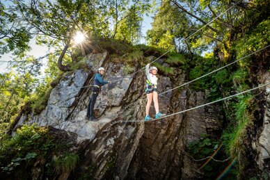 Two kids with helmets and harnesses on rope bridge at rock wall, sunny weather, trees and cliffs in background. | © Snow Space Salzburg, Christoph Huber