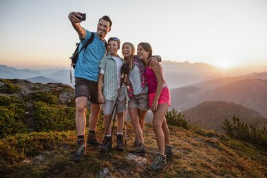Vierköpfige Familie macht Selfie beim Wandern auf dem Berg, Sonnenuntergang hinter Bergsilhouetten im Hintergrund sichtbar