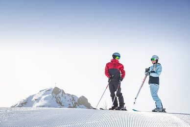 Eine Skifahrerin mit hellblauem Skianzug und ein Skifahrer mit grau-rotem Skianzug stehen lächelnd auf einer frisch präparierten Piste | © Snow Space Salzburg