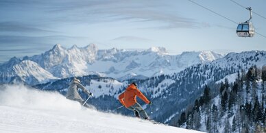 Zwei Skifahrer auf frisch präparierter Piste mit Gondel und Alpenpanorama im Hintergrund