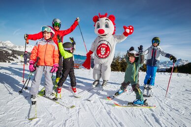 Ein Erwachsener und vier Kindern winken in die Kamera, in der Mitte steht das Maskottchen von Monte Popolo. 