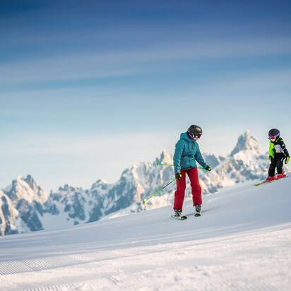 Ein Kind mit fährt mit seinem Papa vor einem Gebirgsstock auf frischpräparierter Piste. | © Snow Space Salzburg