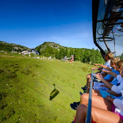 Familie in der Sommer-Sesselbahn zeigt auf Spielplatz und Gamskogelhütte vor Bergkulisse bei Zauchensee.