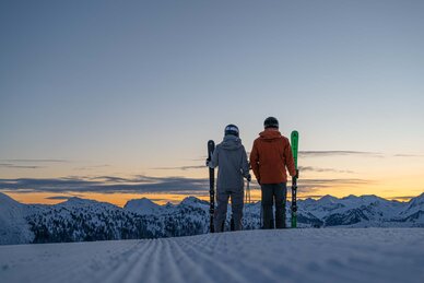 Zwei Skifahrer halten ihre Ski in den Händen und blicken auf den Sonnenaufgang